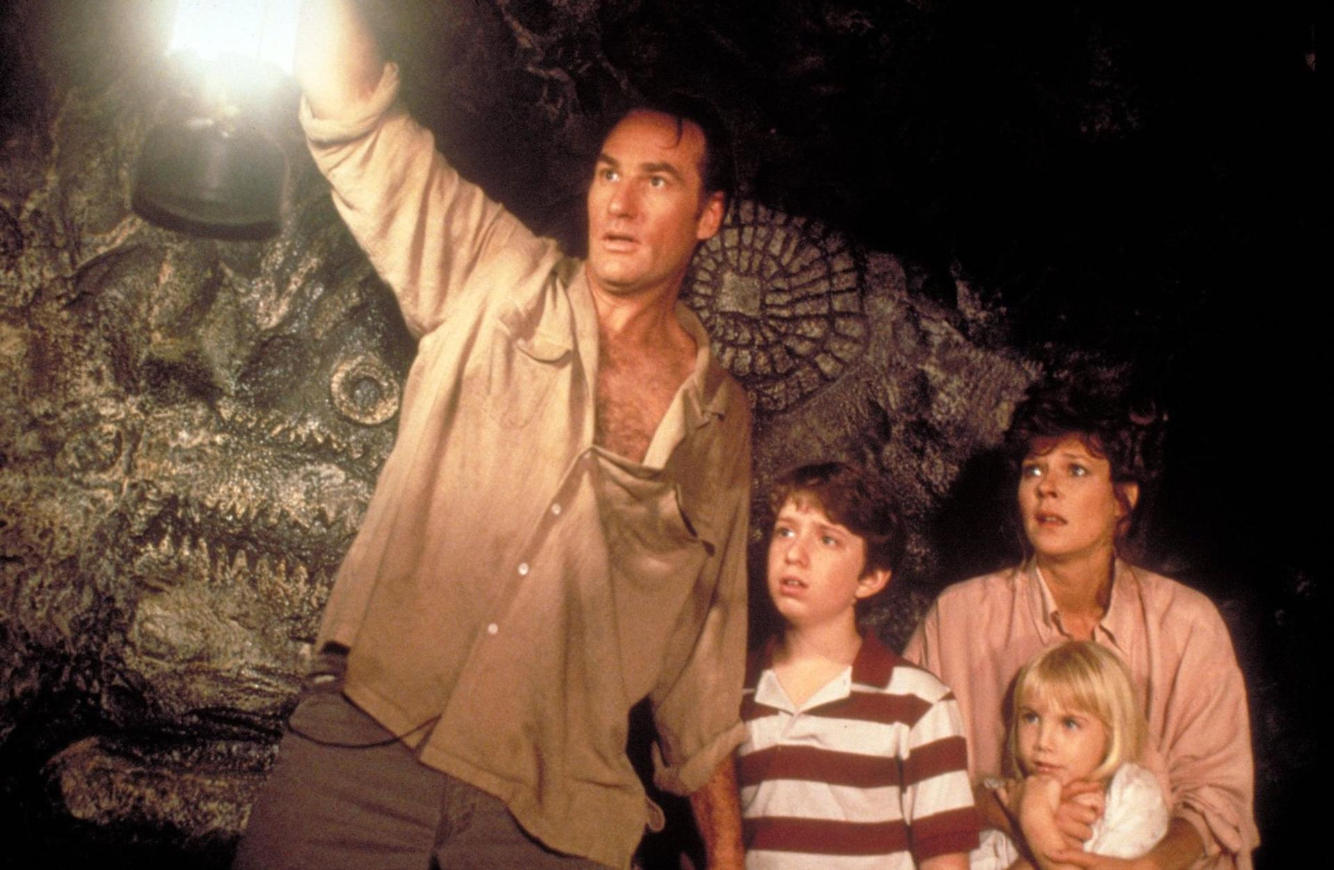 Ein Mann hält eine Lampe in einer dunklen Höhle und beleuchtet eine Familie. Die Familie sieht verängstigt aus. Im Hintergrund befindet sich eine Steinschnitzerei.