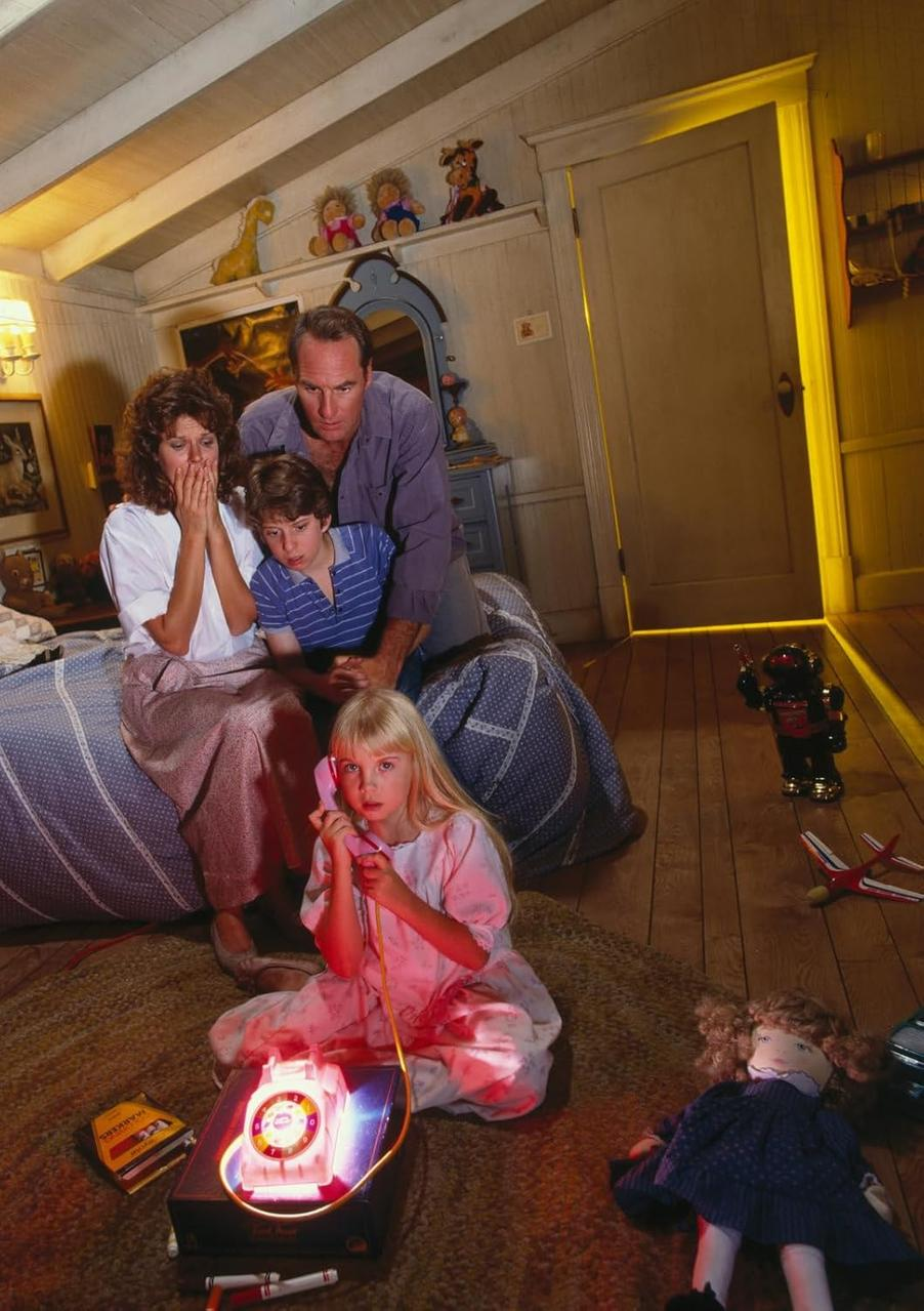 Familie im Schlafzimmer, Mädchen telefoniert. Objekte sind eine Puppe, ein Roboter und ein Flugzeug. Die Familie wirkt besorgt.