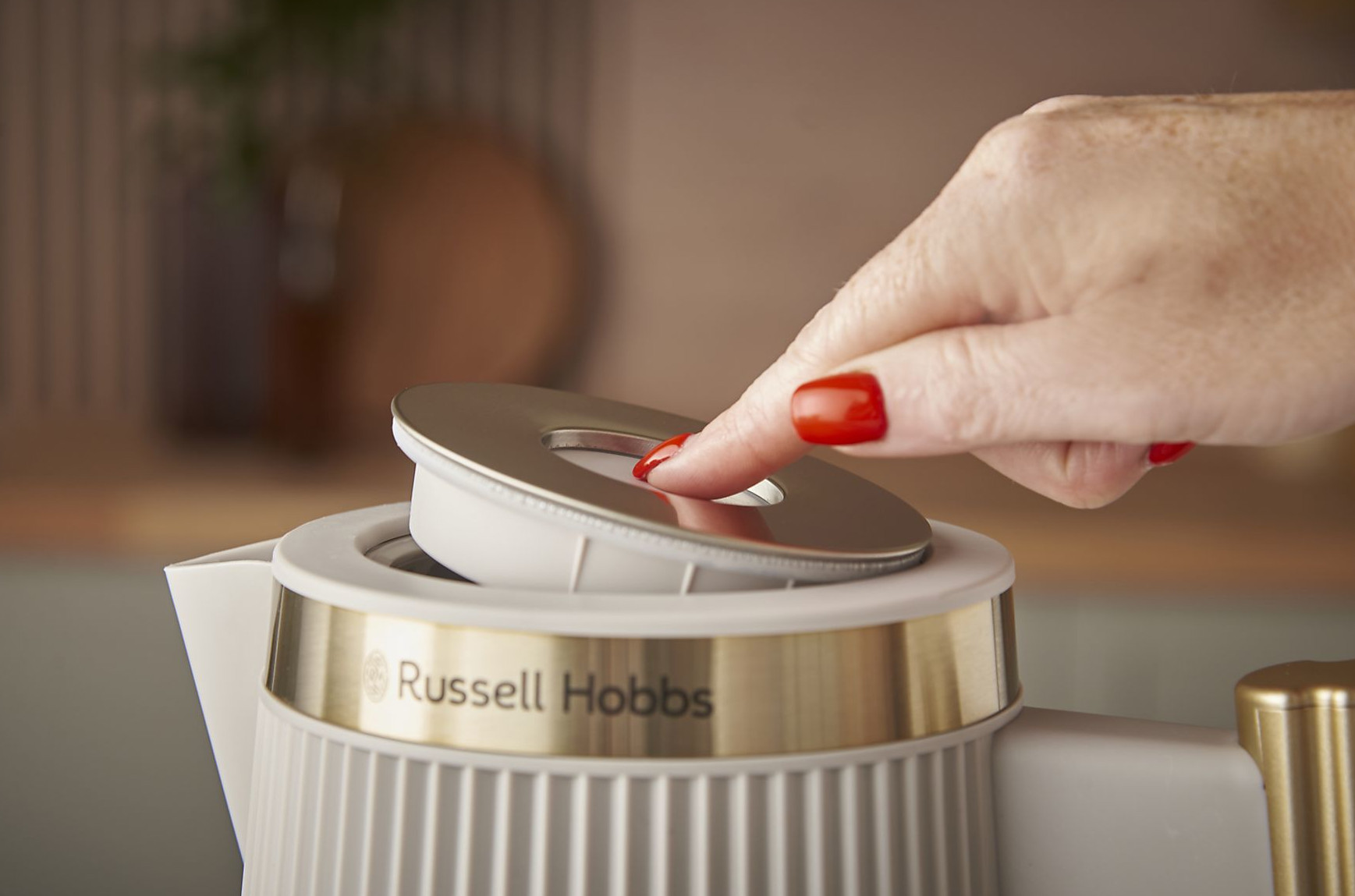 Dłoń z czerwonymi paznokciami naciska pokrywę czajnika Russell Hobbs. Tło jest rozmyte z drewnianą deską.