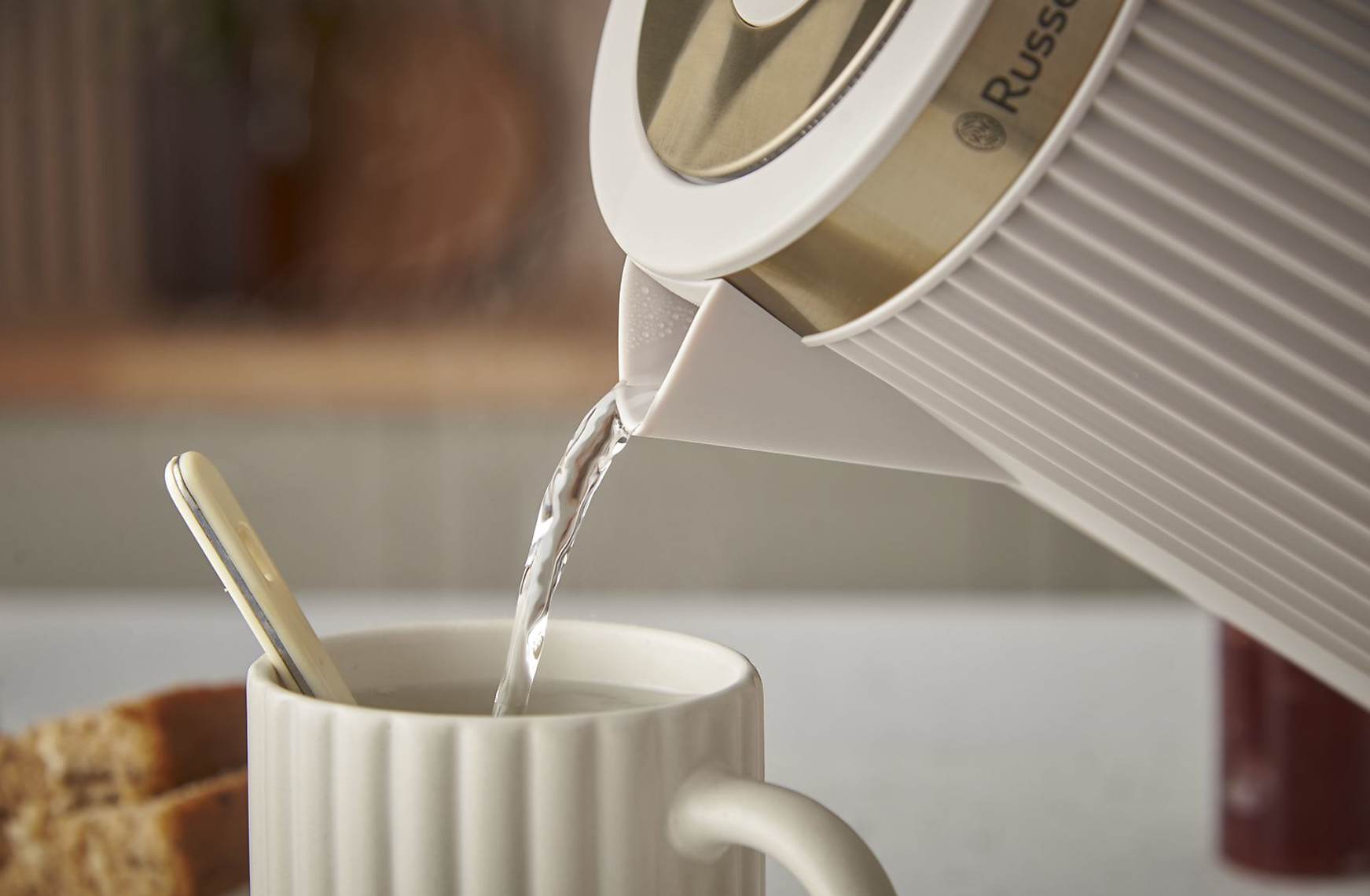 Biały czajnik nalewa wodę do białego kubka z łyżeczką. Na stole leży pokrojony chleb.