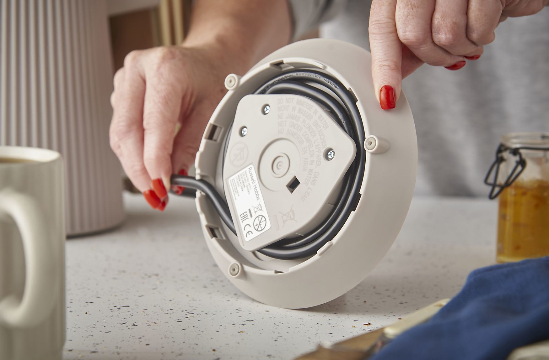 Osoba trzyma białą podstawę czajnika z czarnym przewodem. Widoczne są czerwone paznokcie. Inne przedmioty są w tle.