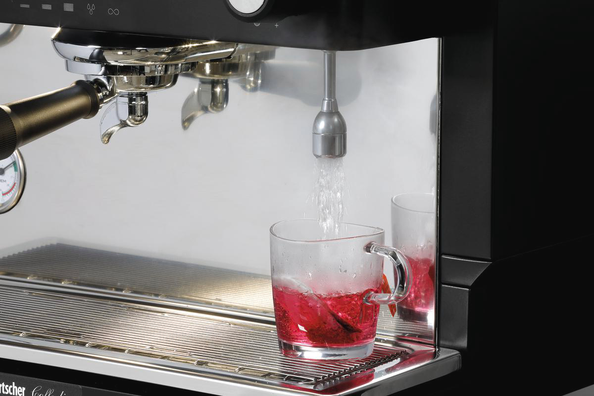 Eine Espressomaschine gibt Wasser in eine gläserne Tasse mit roter Flüssigkeit ab. Dampf steigt auf. Daneben steht eine zweite Tasse mit roter Flüssigkeit.