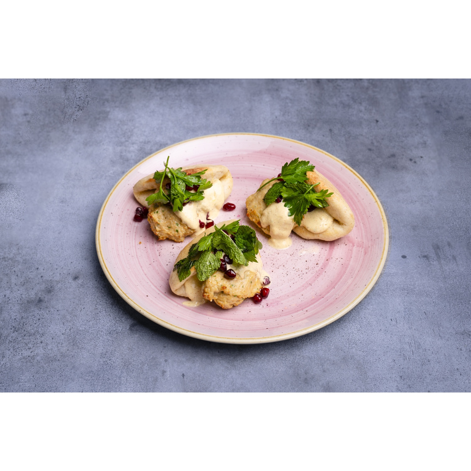 Tre pasticcini ripieni su un piatto rosa. Sono ricoperti di salsa cremosa, erbe aromatiche e semi di melograno. Lo sfondo è grigio.