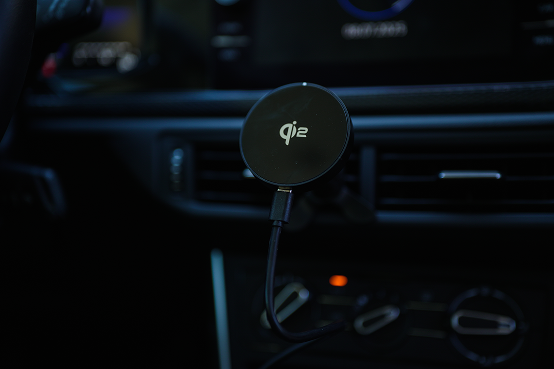 Egy fekete vezeték nélküli töltő USB-kábellel egy autóba szerelve. A töltőn a qi2 logó látható. A háttér elmosódott.