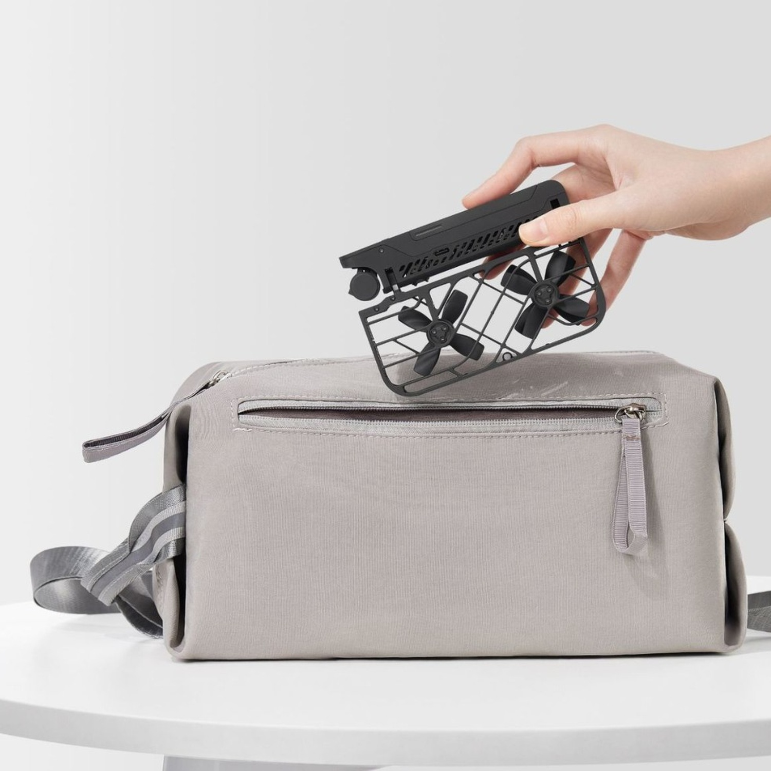 Una mano tiene un drone nero sopra una borsa grigia. Il drone ha delle eliche. La borsa è su un tavolo bianco.