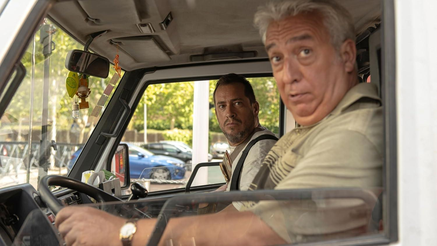 Zwei Männer in einem Auto. Einer fährt, der andere schaut in die Kamera. Bäume und Autos sind durch die Fenster zu sehen.