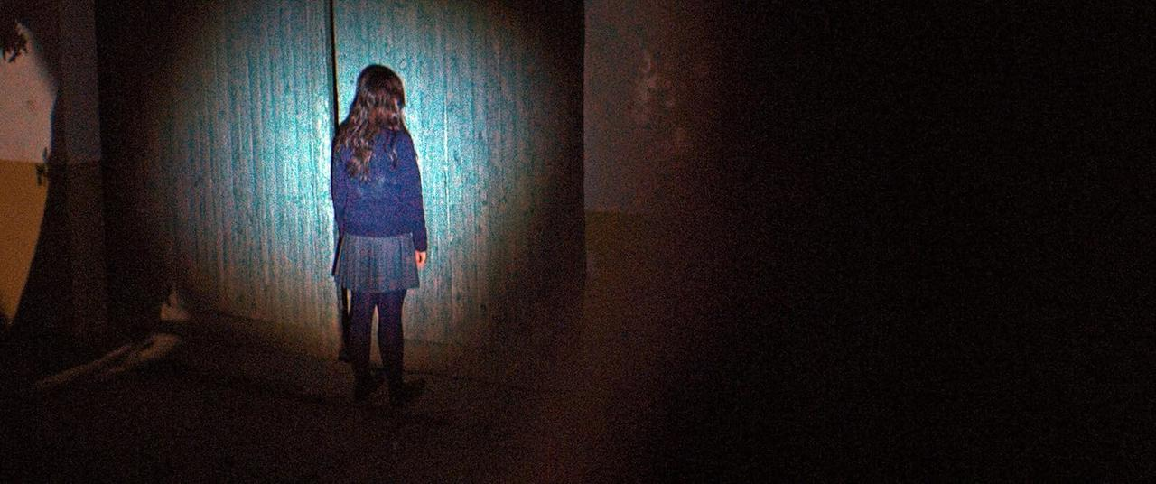 Ein Mädchen steht in einem schwach beleuchteten Raum vor einer Wand. Sie trägt einen blauen Pullover und einen karierten Rock. Die Wand ist türkis.