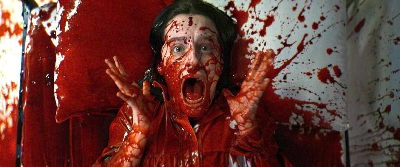 Eine Frau schreit, blutbedeckt. Rote Spritzer auf weißem Hintergrund. Sie hat geweitete Augen und offenen Mund, Hände erhoben.
