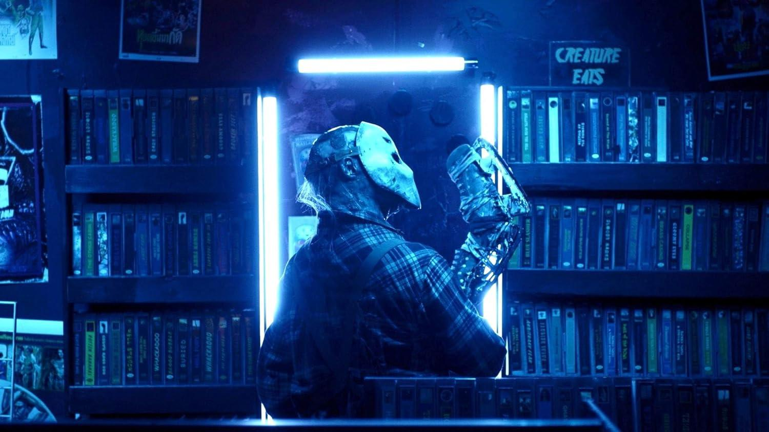 Eine Person mit Maske und kariertem Hemd steht in einem Raum mit VHS-Kassetten. Der Raum ist schwach beleuchtet, mit einem Schild mit der Aufschrift 'CREATURE EATS'.