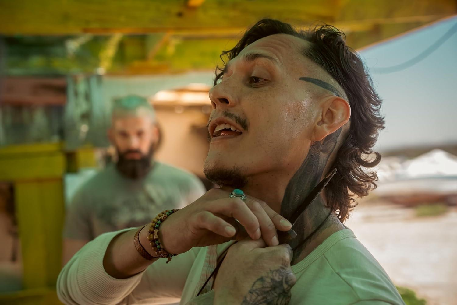 Mann mit Tattoos, Schnurrbart und Ring, hält etwas an seinen Hals. Ein anderer Mann ist im Hintergrund unscharf.