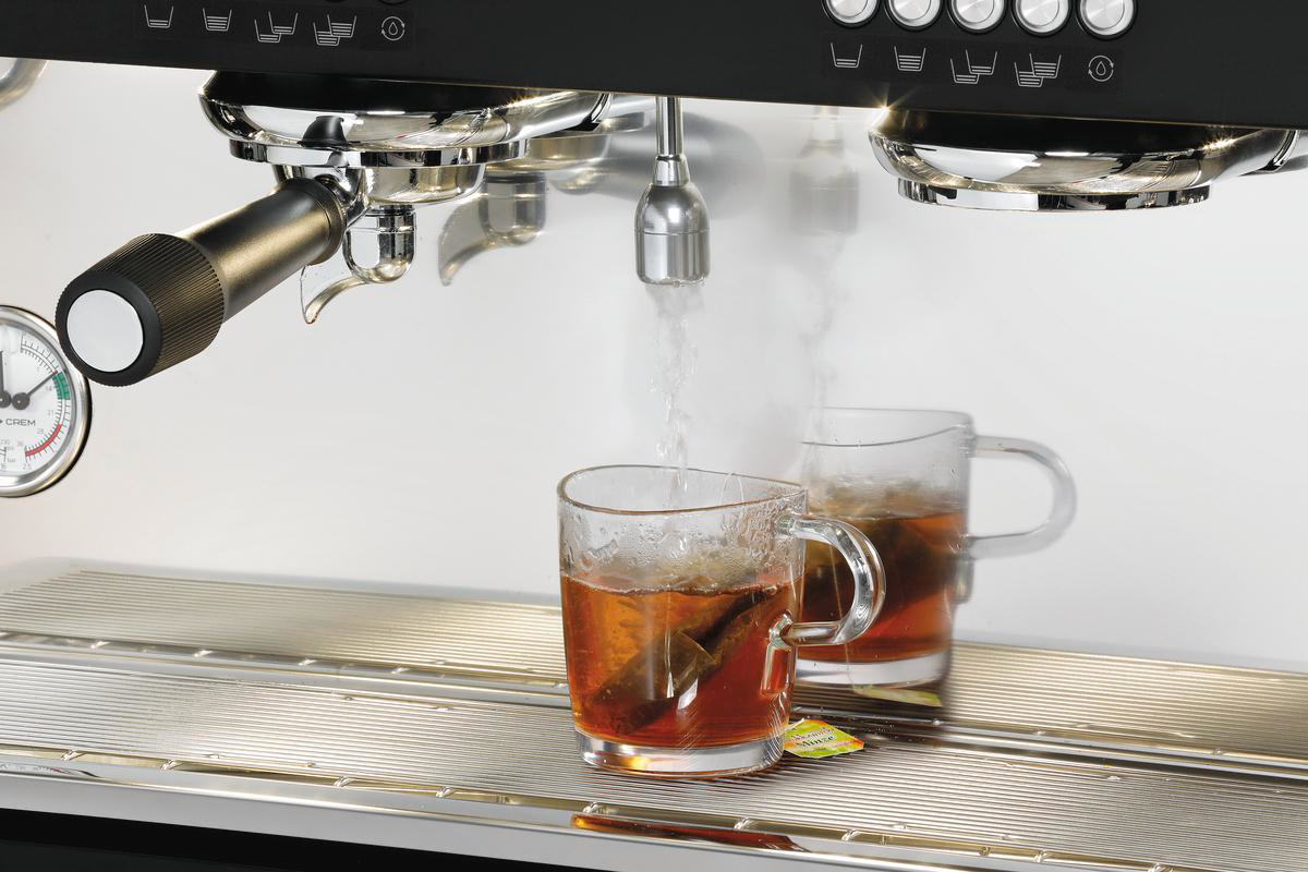 Tee wird gebrüht. Wasser gießt in zwei Glasbecher mit Teebeuteln. Eine Espressomaschine befindet sich oben. Die Einstellung ist eine Küche.