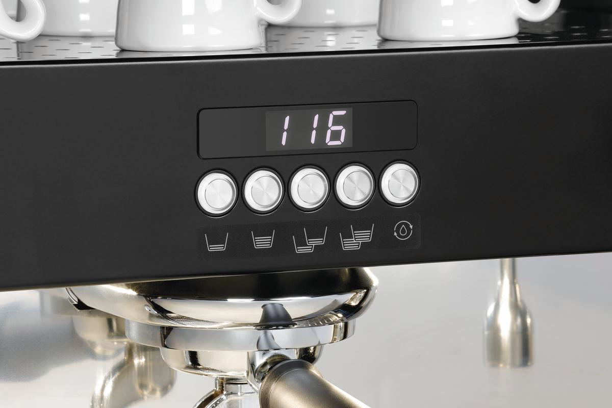 Nahaufnahme einer schwarzen Kaffeemaschinen-Bedienblende. Sie zeigt '116' und hat Knöpfe. Oben sind weiße Kaffeetassen.