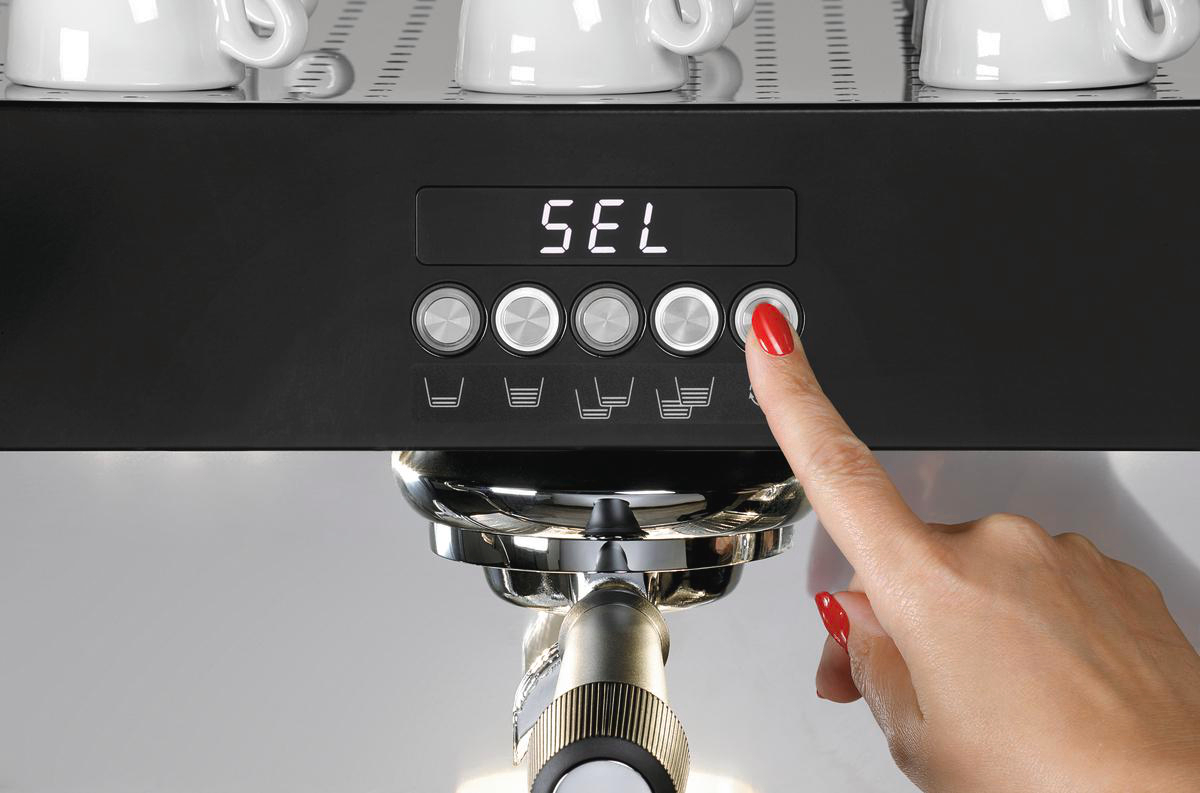 Eine Hand mit rotem Nagellack drückt eine Taste an einer Kaffeemaschine. Die Maschine zeigt 'SEL' an und hat weitere Knöpfe.