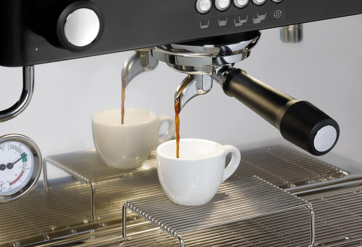 Espressomaschine gießt Kaffee in zwei weiße Tassen auf einem Metallgitter. Schwarze Maschine mit Chromakzenten, Manometer links.