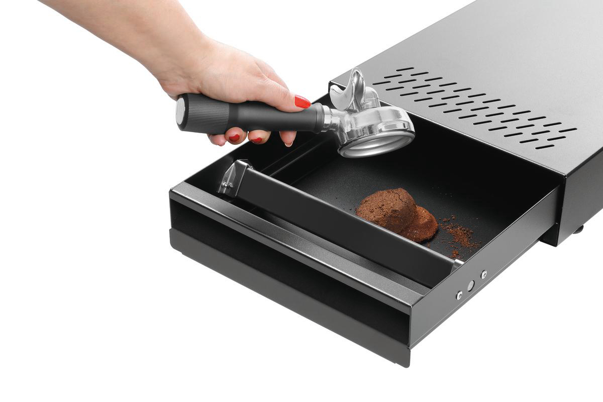 Eine Hand hält einen Siebträger über einer Schublade. Die Schublade enthält gebrauchten Kaffeesatz. Die Espressomaschine ist silber und schwarz.