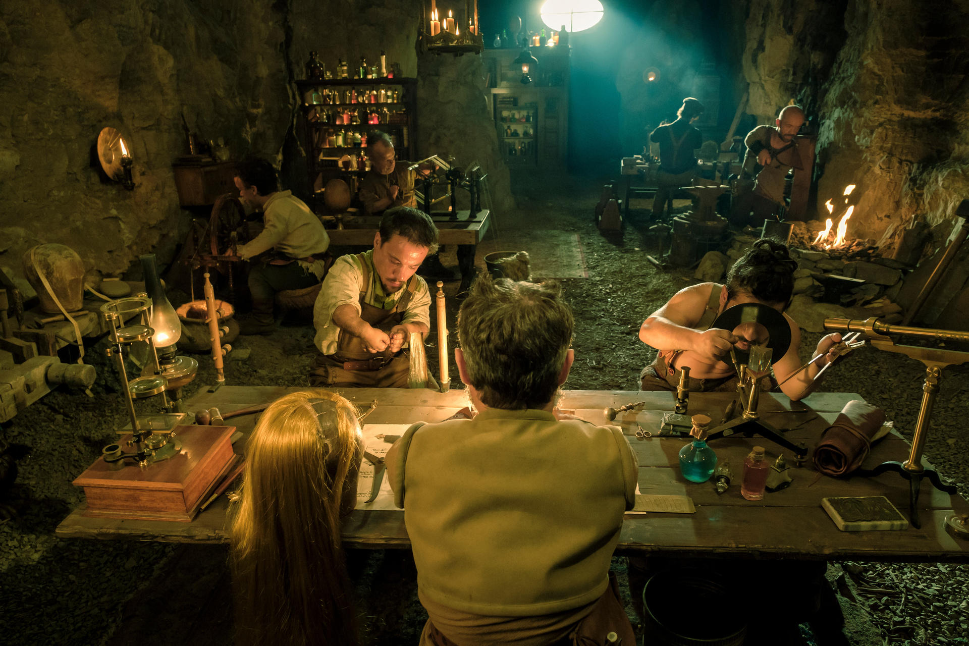 In einer Höhle arbeiten mehrere Personen an einem Tisch. Kerzen und Lampen spenden Licht. Werkzeuge und Materialien sind verstreut.