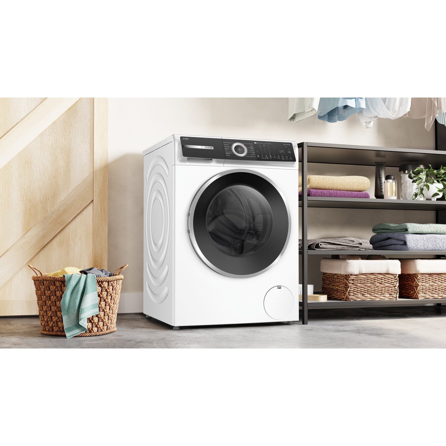 Een witte wasmachine staat naast een houten deur en een plank met handdoeken. Er staat een mand met kleren voor.