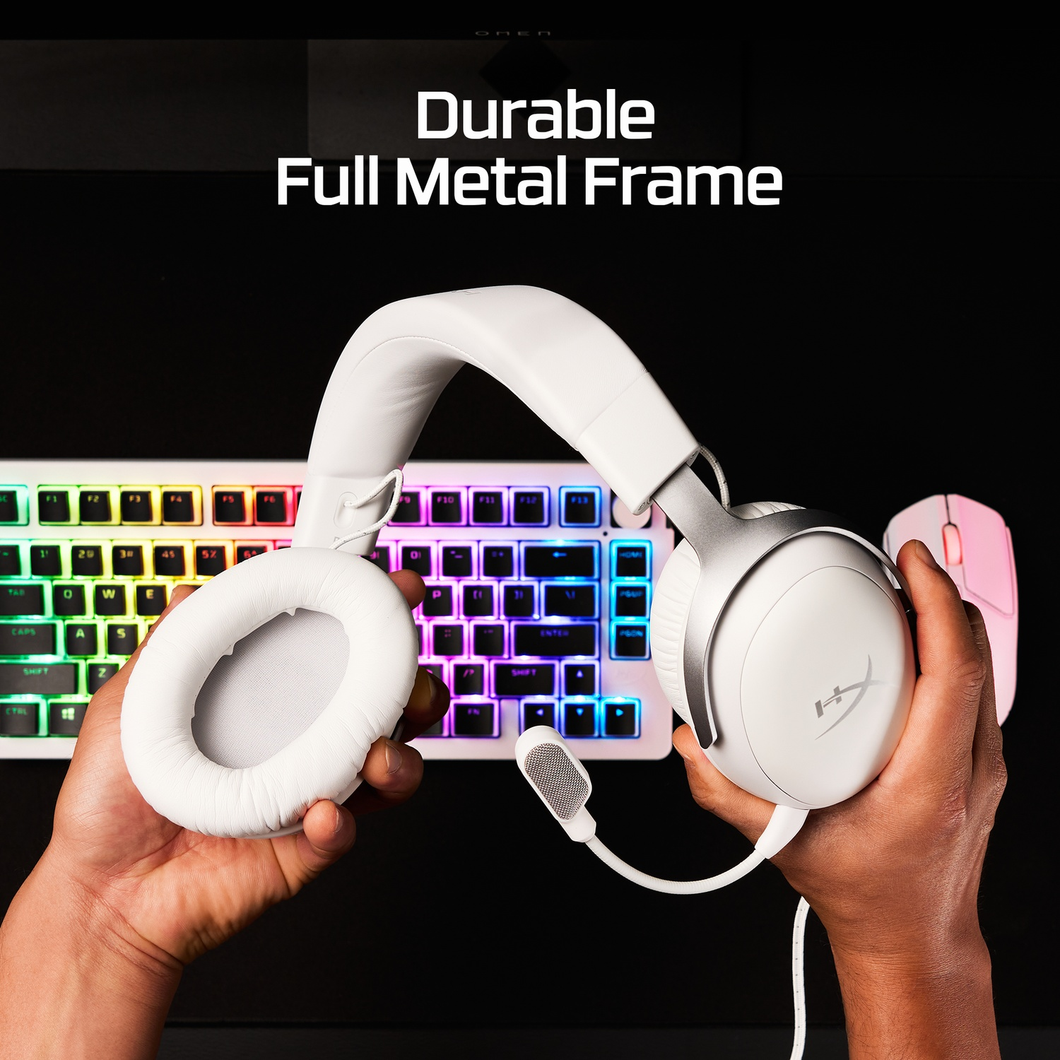 A person holds white headphones with a microphone. Behind is a keyboard with rainbow lights and a pink mouse.