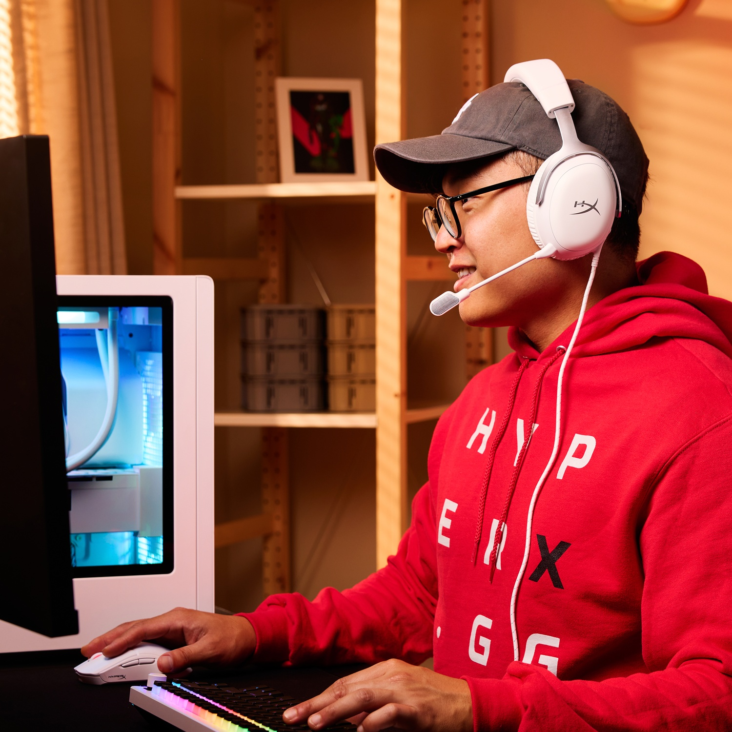 A man wearing a headset and glasses is gaming. He's wearing a red hoodie, cap, and is sitting in front of a computer.