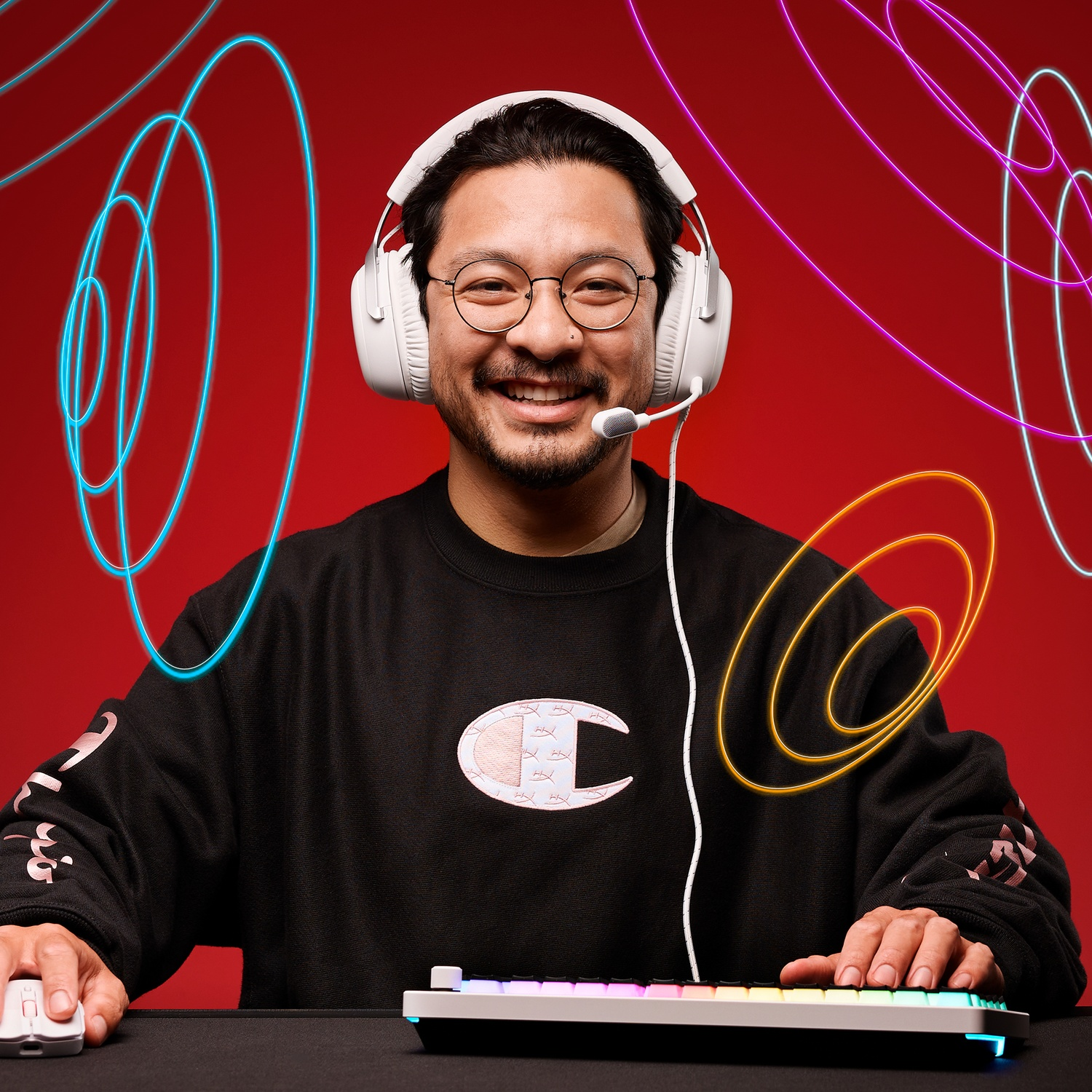 Smiling man with glasses and headset, using a keyboard and mouse. He wears a black sweatshirt against a red backdrop with neon lines.