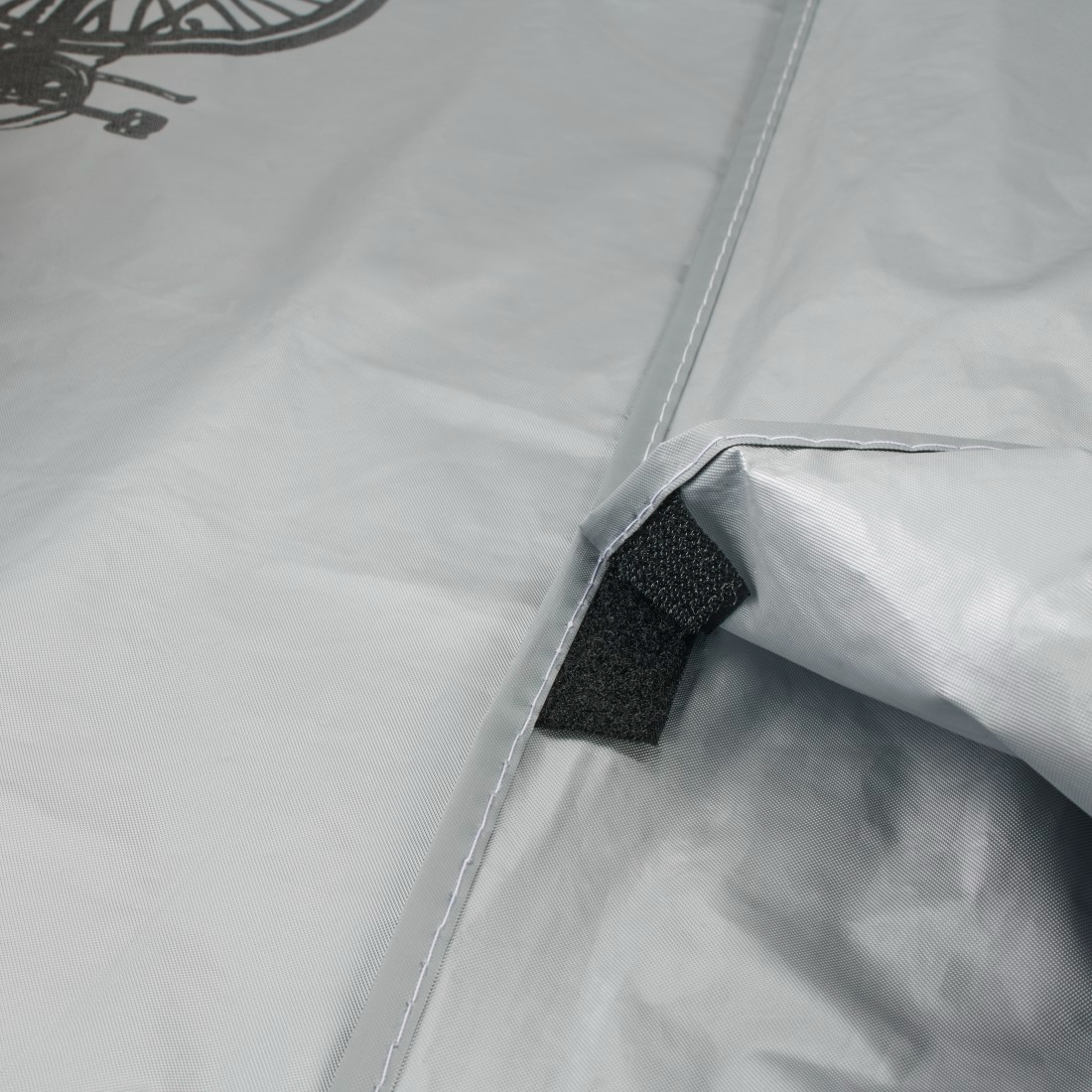 Közelkép egy világosszürke, feltehetően vízálló anyagról. Egy fekete tépőzáras pánt látható. Bal felül egy kerékpár illusztráció.