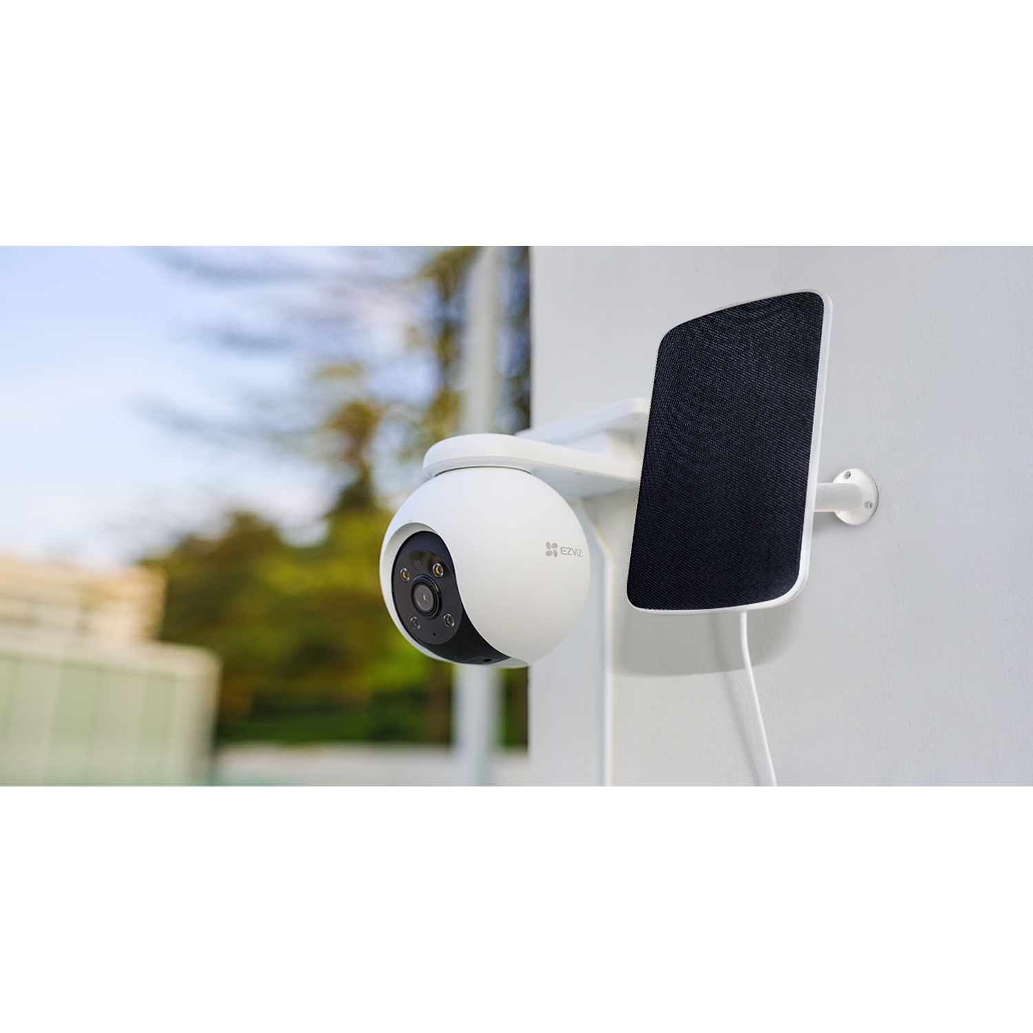 Una telecamera di sicurezza bianca con un altoparlante nero è montata su un muro bianco. Lo sfondo mostra alberi e un cielo sfocato.
