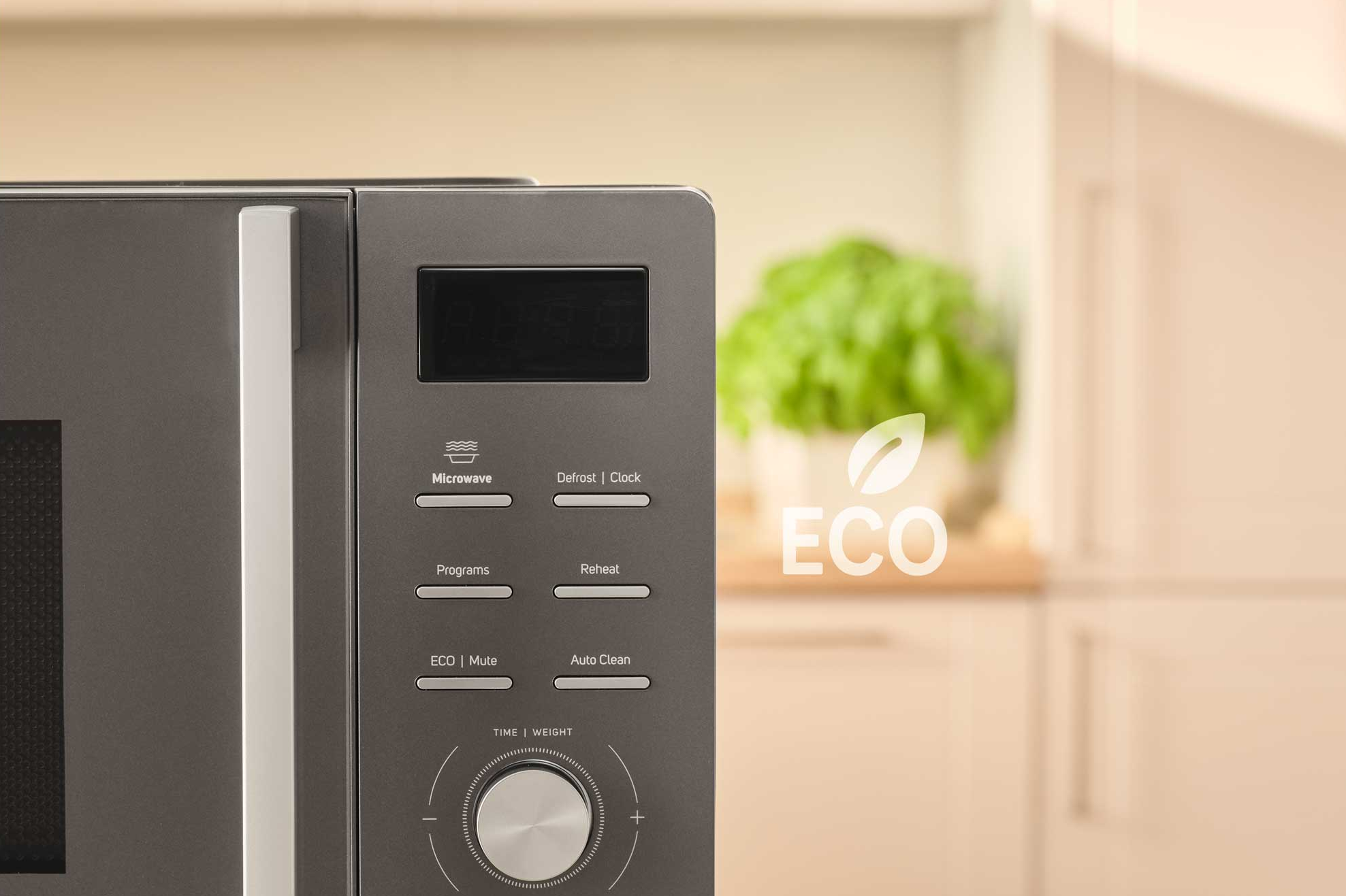 Egy szürke mikrohullámú sütő vezérlőgombokkal, digitális kijelzővel és tárcsával. A háttérben egy zöld növény és az ECO szöveg látható.