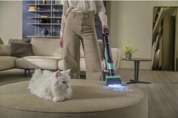 Una persona aspira un pouf rotondo con un gatto bianco. L'aspirapolvere è verde acqua e nero. Un divano e delle mensole sono sullo sfondo.