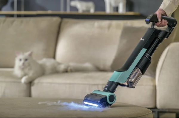 Una persona aspira un divano con un aspirapolvere moderno. Un gatto bianco riposa sul divano. La polvere è visibile.