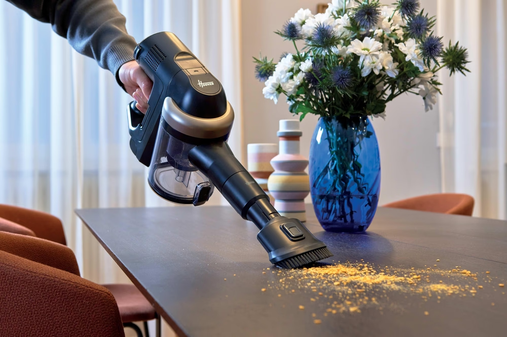 Una persona aspira un tavolo scuro con un Hoover nero e argento. Un vaso blu con fiori e vasi decorativi sono sullo sfondo.