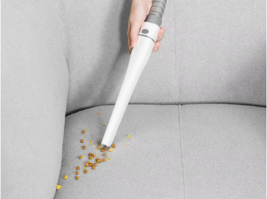 Une main aspire des miettes d'un canapé gris. L'aspirateur est blanc, avec un tuyau gris. Les miettes sont marron et jaunes.
