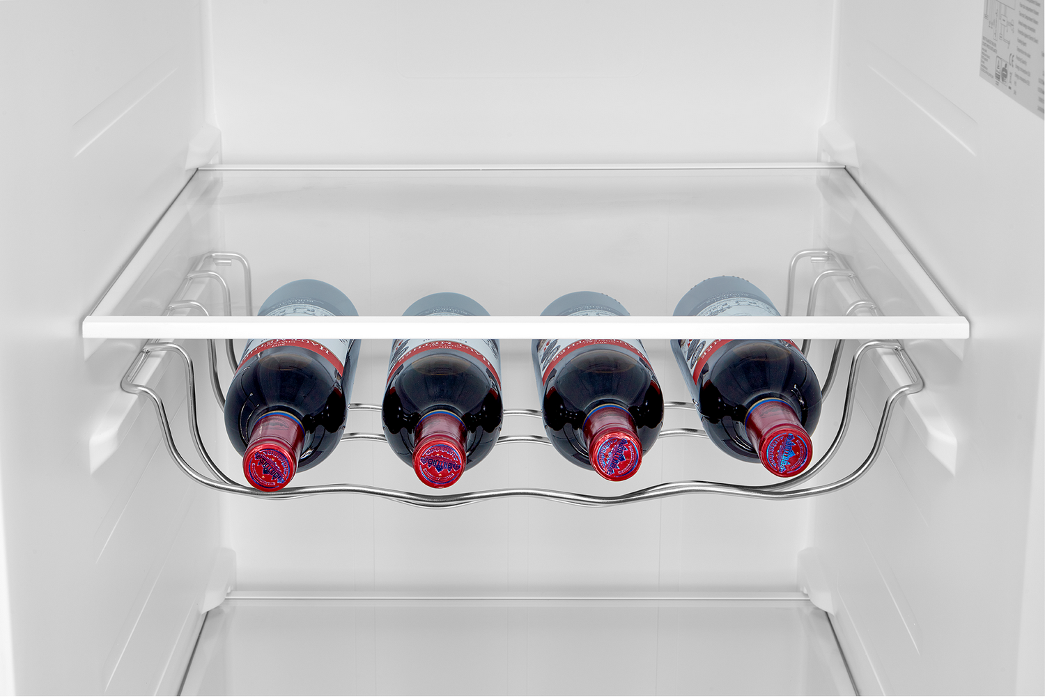 Dentro de un refrigerador, cuatro botellas de vino están en una rejilla metálica. Las botellas tienen tapas rojas y líquido oscuro, en un interior blanco.