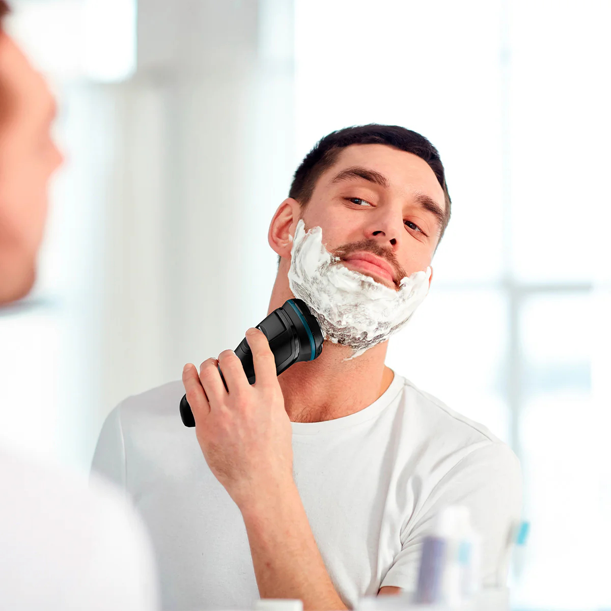 Un hombre se afeita la cara con una afeitadora eléctrica frente a un espejo. Tiene crema de afeitar en la barba y el cuello.