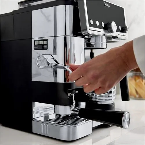 Una mano opera una máquina de espresso Ninja. La máquina es negra y cromada, la mano gira una manija plateada.