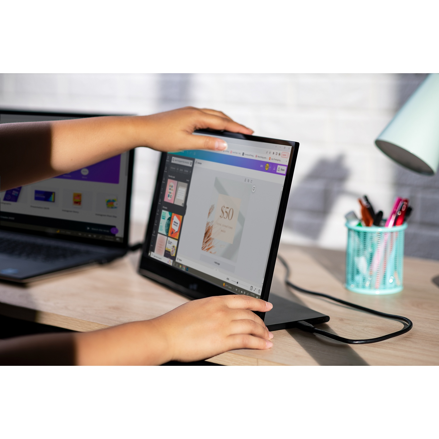 Hands near laptop, showing a design interface. A second laptop and desk items are also present. The scene is lit by sunlight.