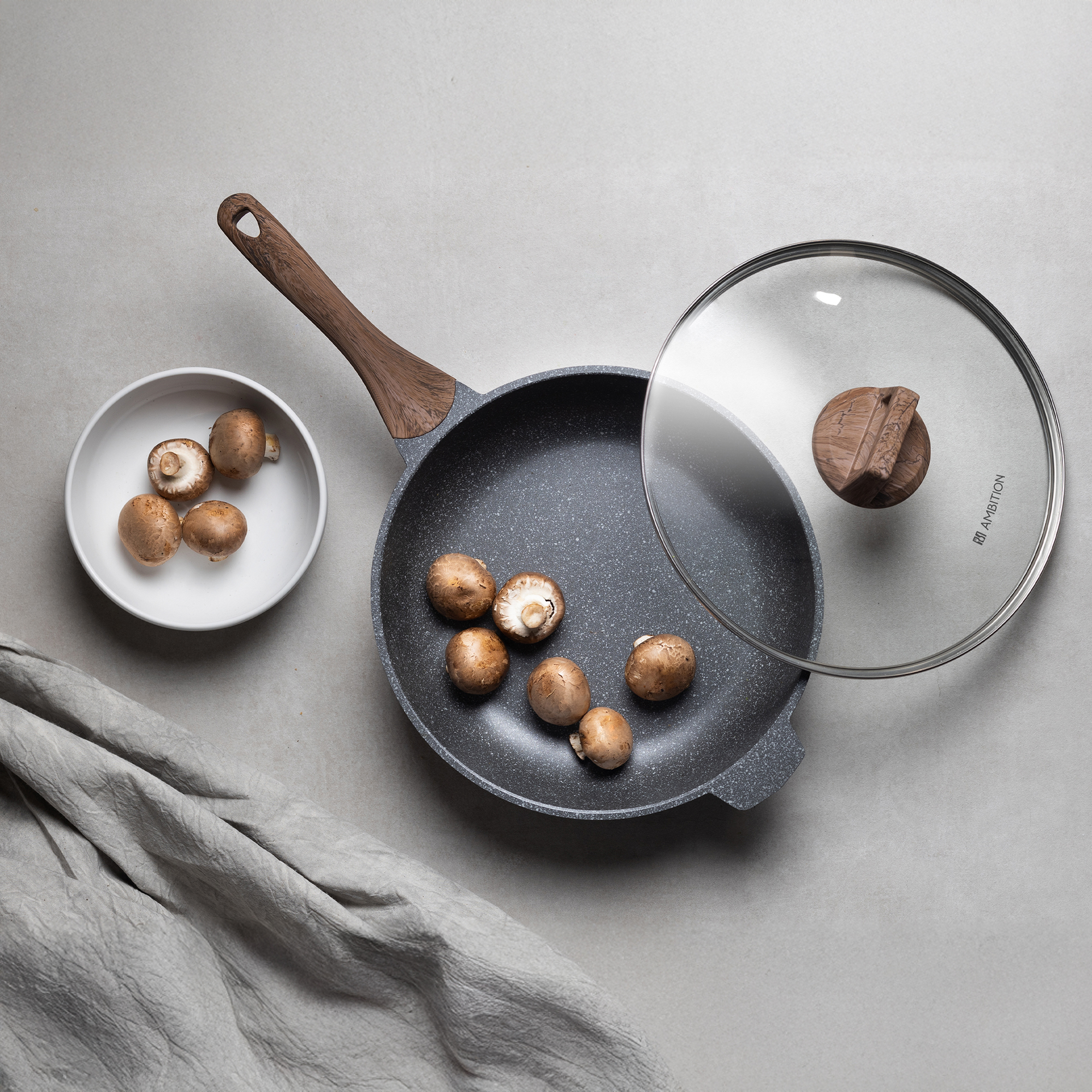 Widok z góry na patelnię, pokrywkę i miskę z grzybami. Brązowe grzyby są na patelni, w misce i na stole. Po lewej stronie jest ściereczka.