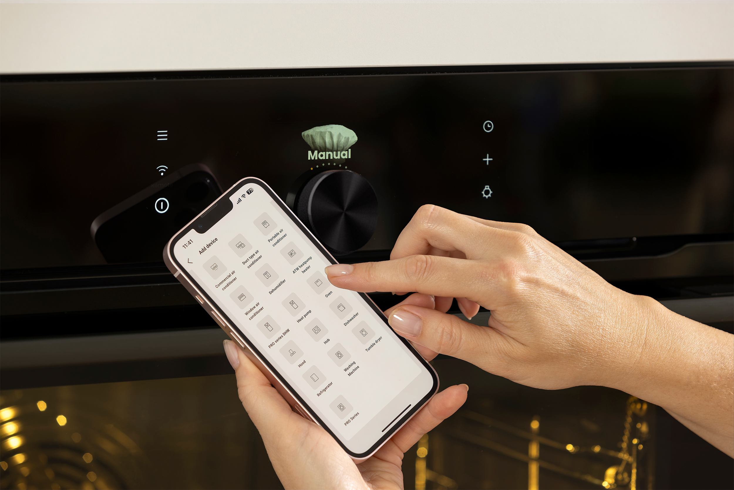 A hand holds a phone over an oven. The phone shows device options. The oven has a manual control button.