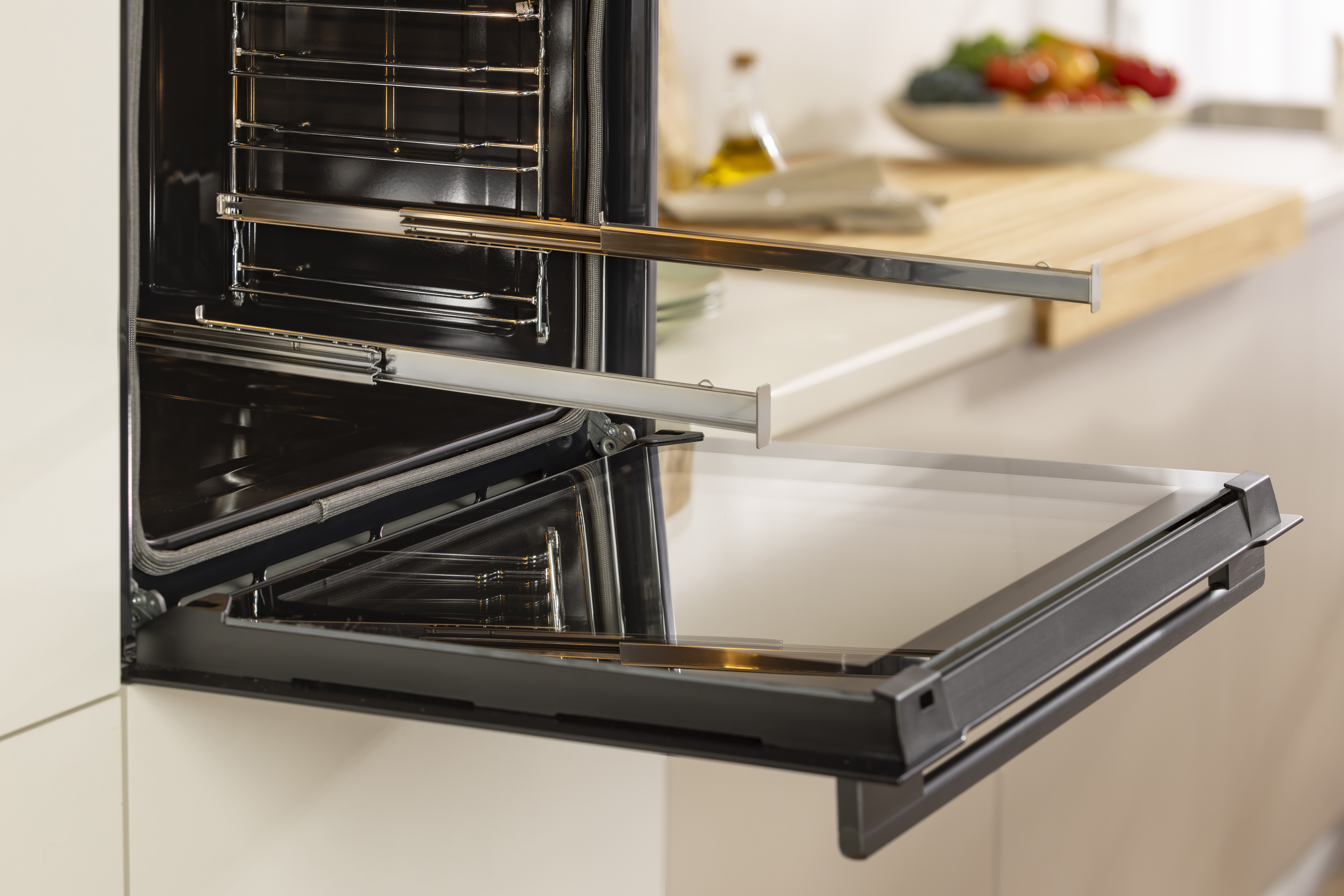 Open oven showing racks, glass door, and interior. Background: kitchen counter with vegetables on a wooden board.
