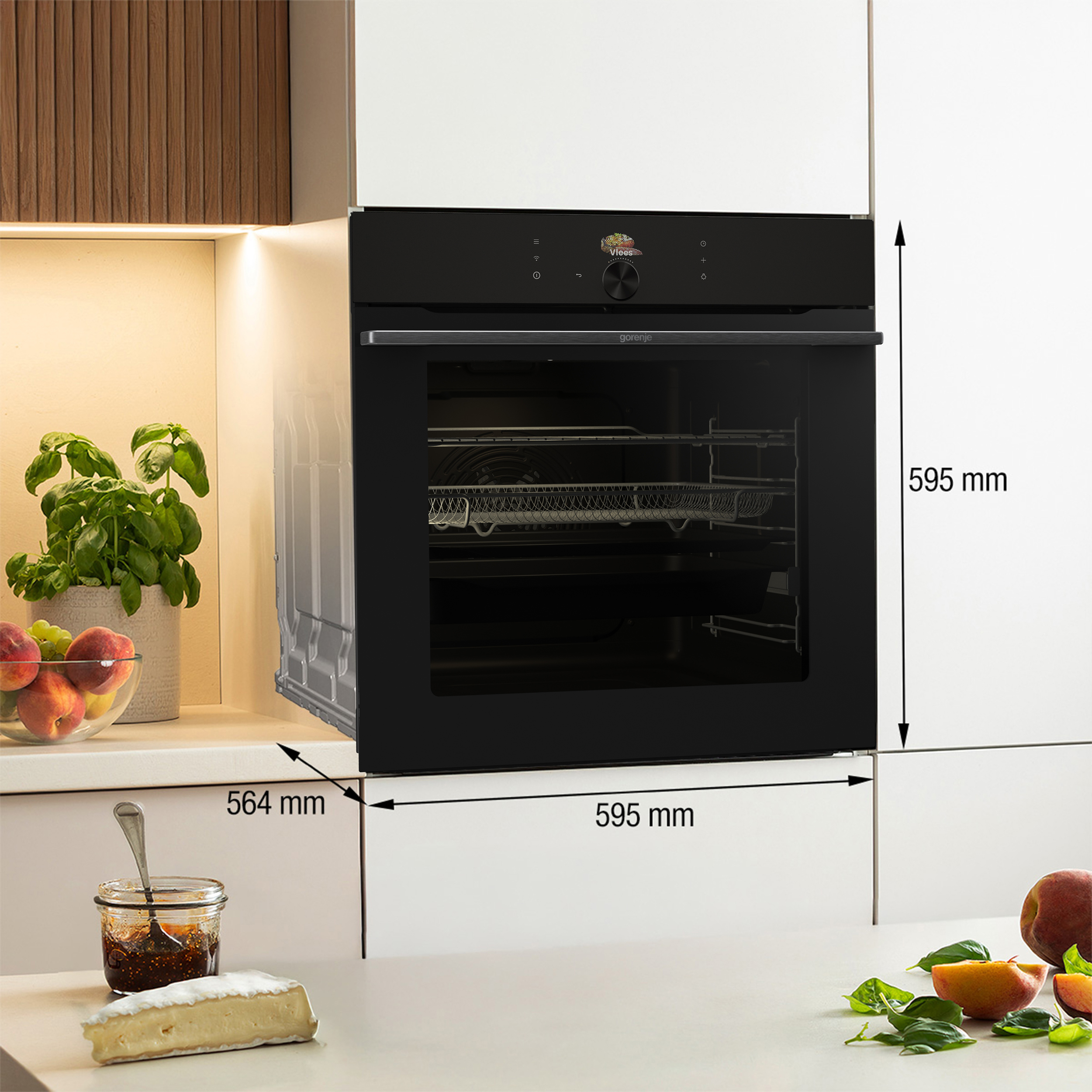 A black oven in a white kitchen, with dimensions. On the counter, fruits, basil, cheese, and jam are visible.