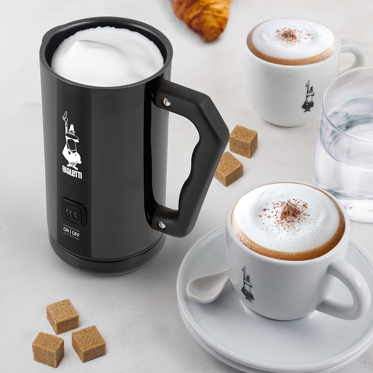 Un montalatte nero, cappuccini, zollette di zucchero e un croissant su una superficie bianca. Tazze con logo Bialetti.