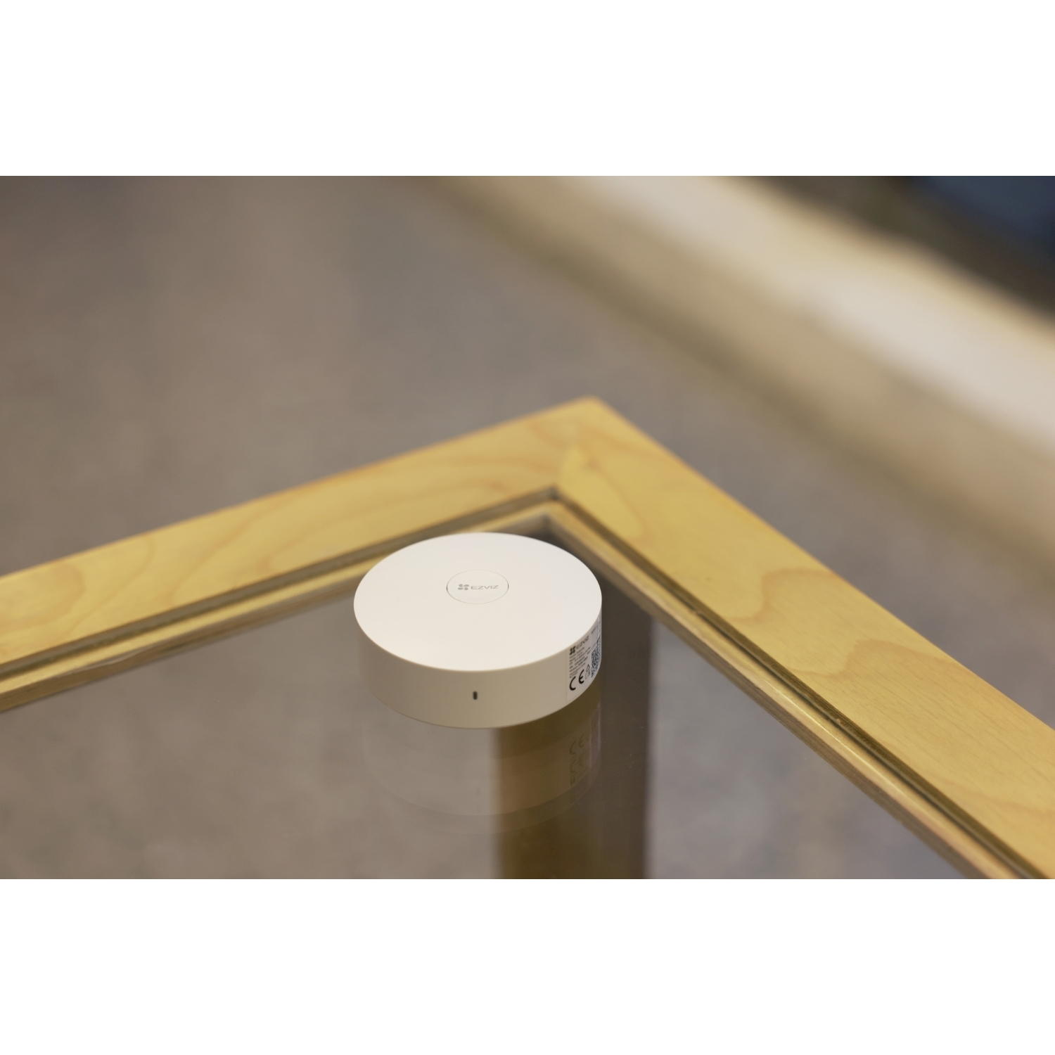 Un dispositivo bianco e rotondo con logo EZVIZ è su una superficie di vetro. Una cornice in legno chiaro circonda il vetro. Sfondo sfocato.
