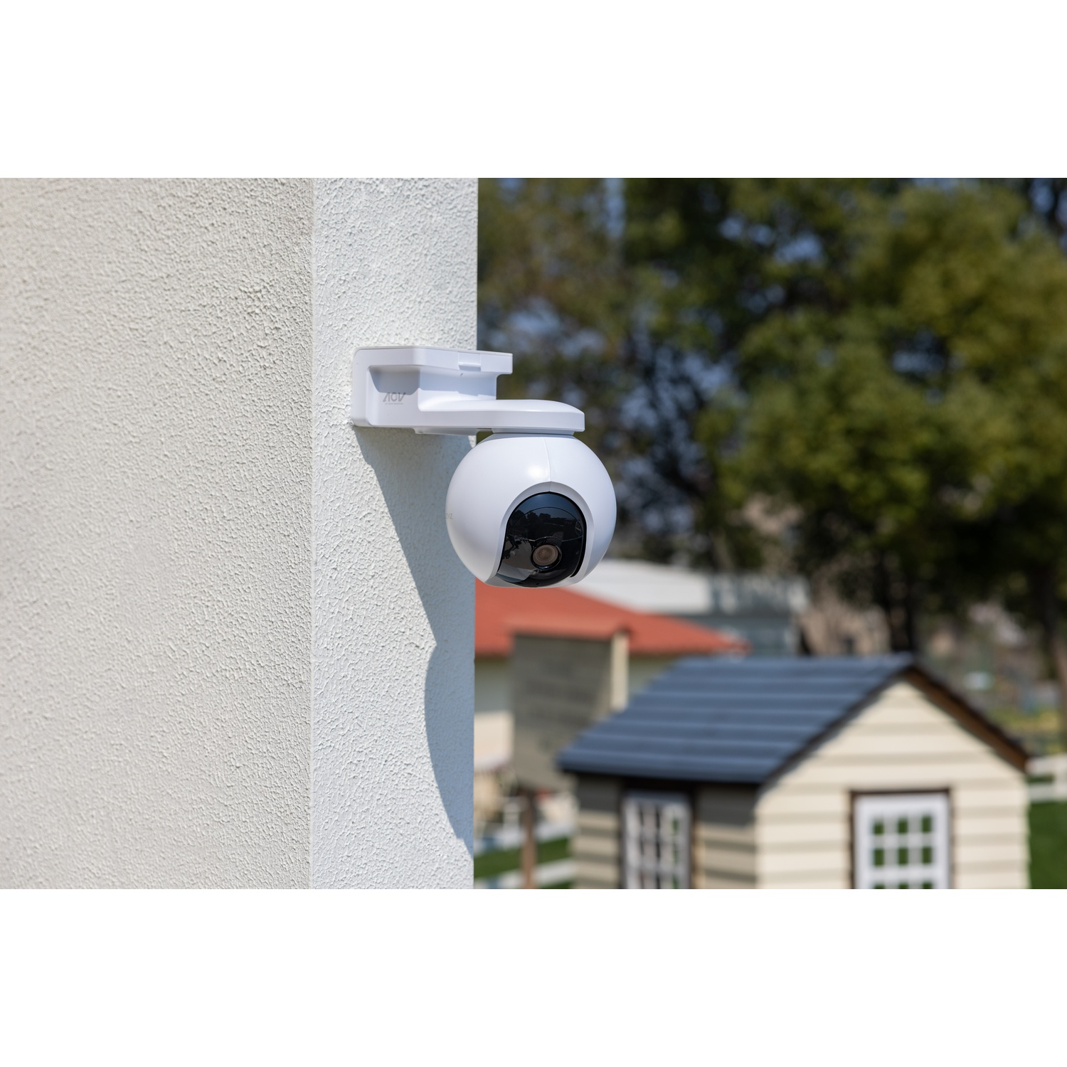 Una telecamera di sicurezza bianca montata su un muro strutturato. Lo sfondo mostra un giardino sfocato con una casetta e alberi.