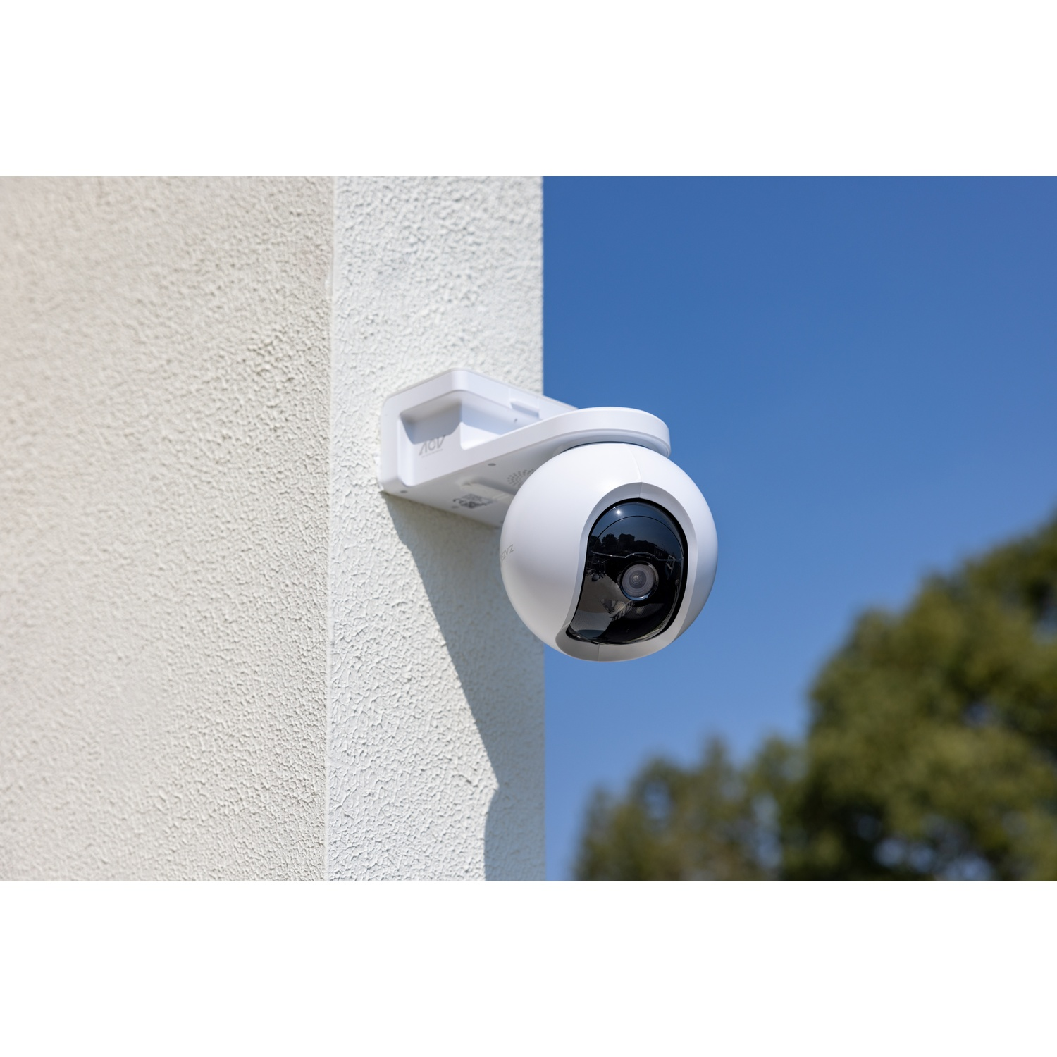 Una telecamera di sicurezza bianca montata su un muro strutturato contro un cielo blu. Gli alberi sono visibili sullo sfondo.