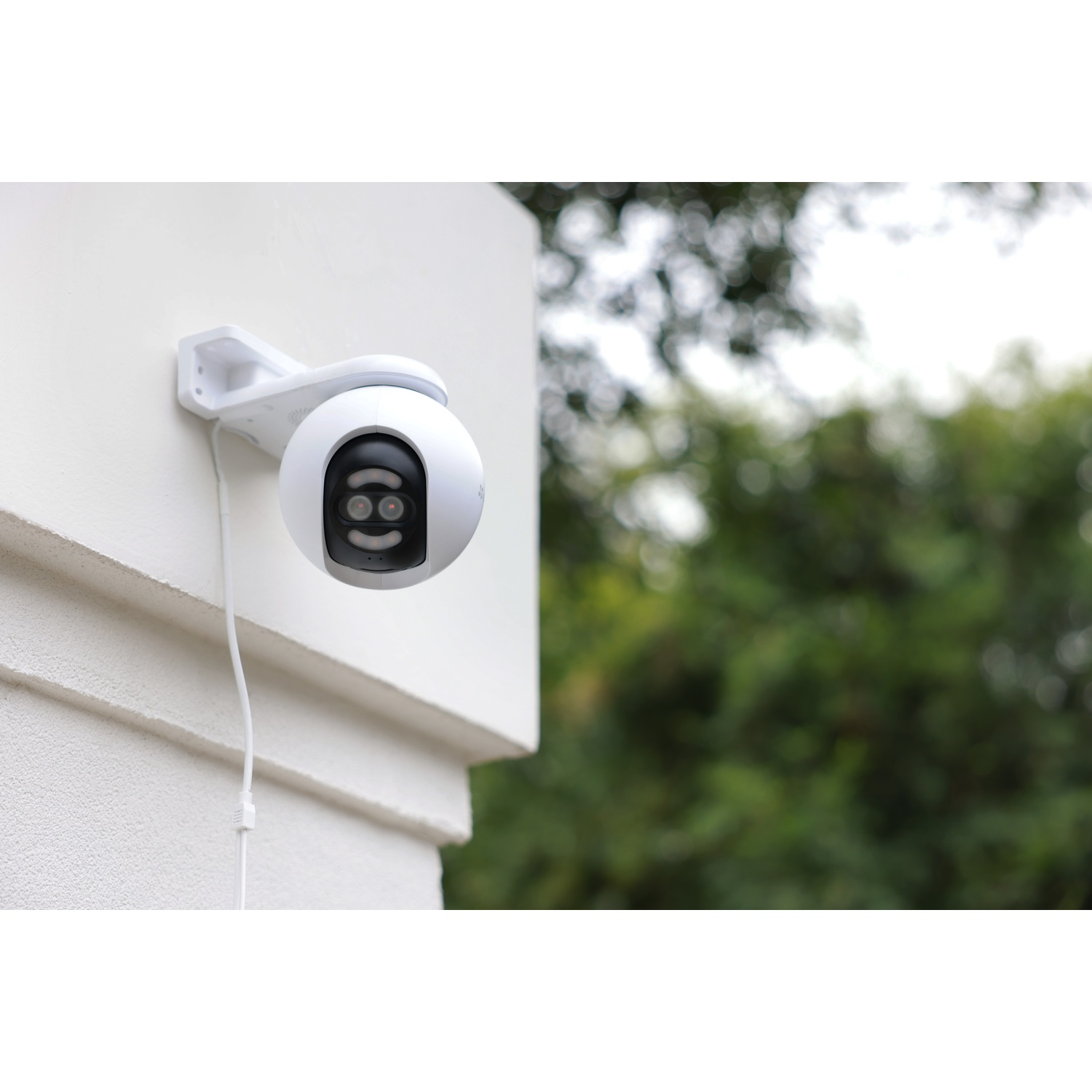 Una telecamera di sicurezza bianca montata su un muro bianco. Fogliame verde sullo sfondo sfocato. La telecamera è rotonda.