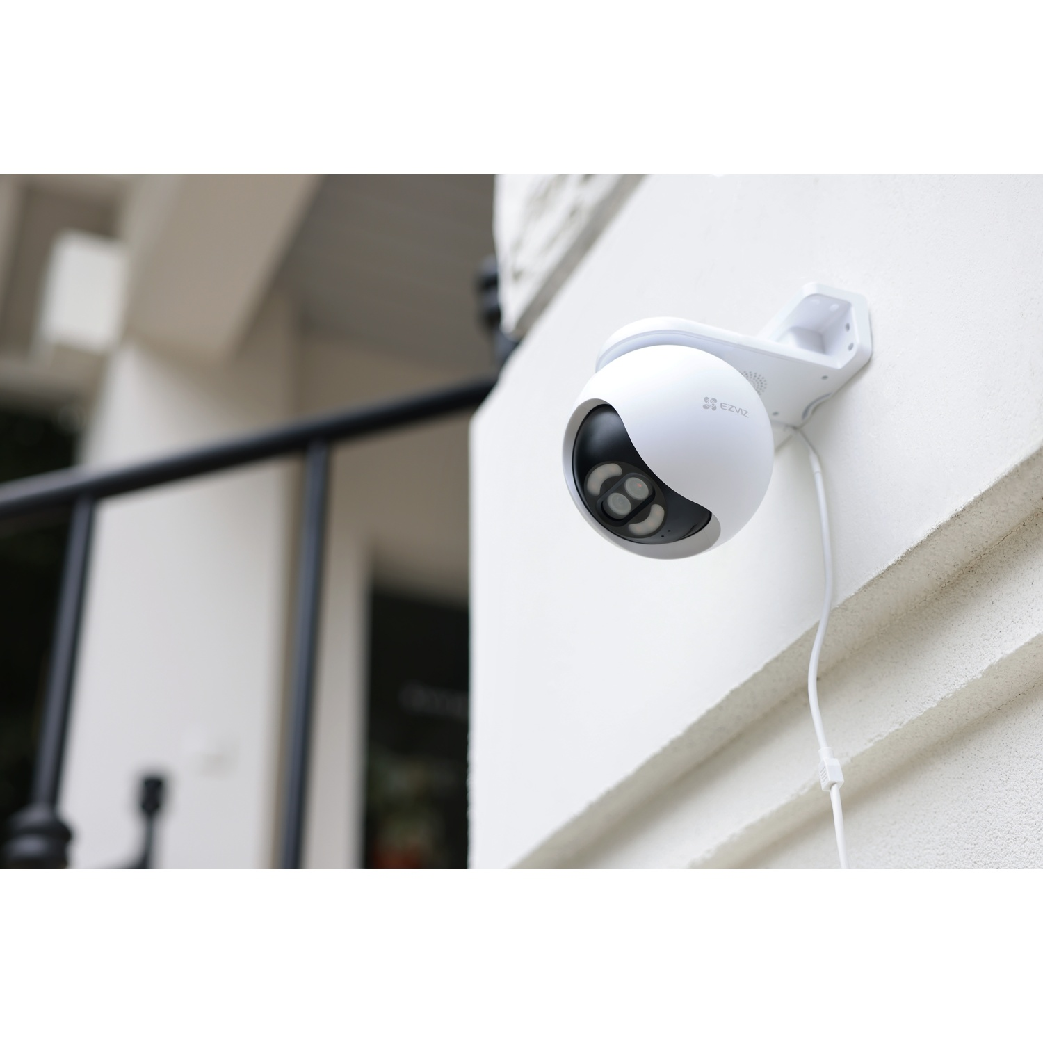 Una telecamera di sicurezza bianca montata su un muro bianco. Dettagli neri sulla telecamera e un cavo. Ringhiera nera sullo sfondo.