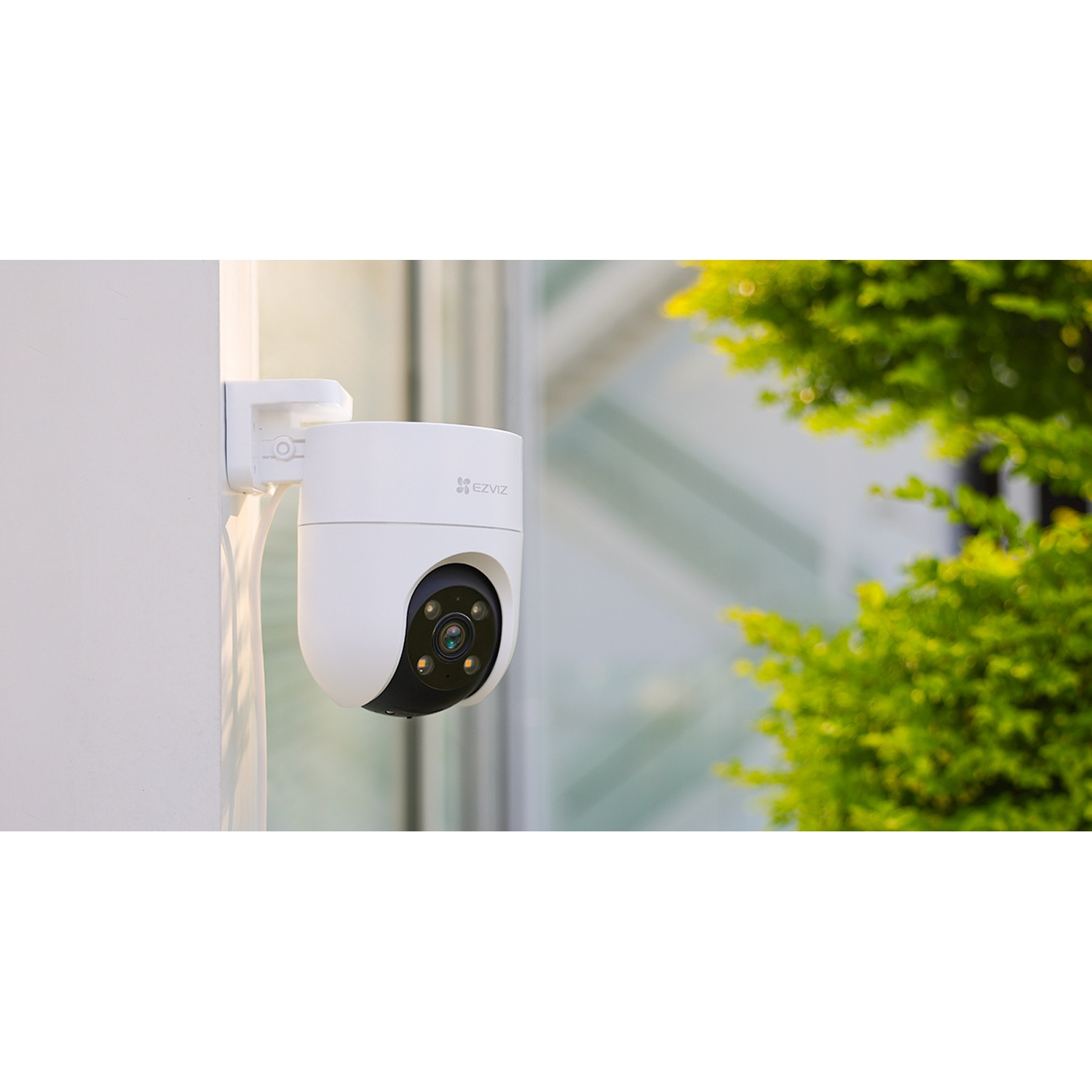 Una telecamera di sicurezza EZVIZ bianca montata su un muro bianco. Fogliame verde sullo sfondo.