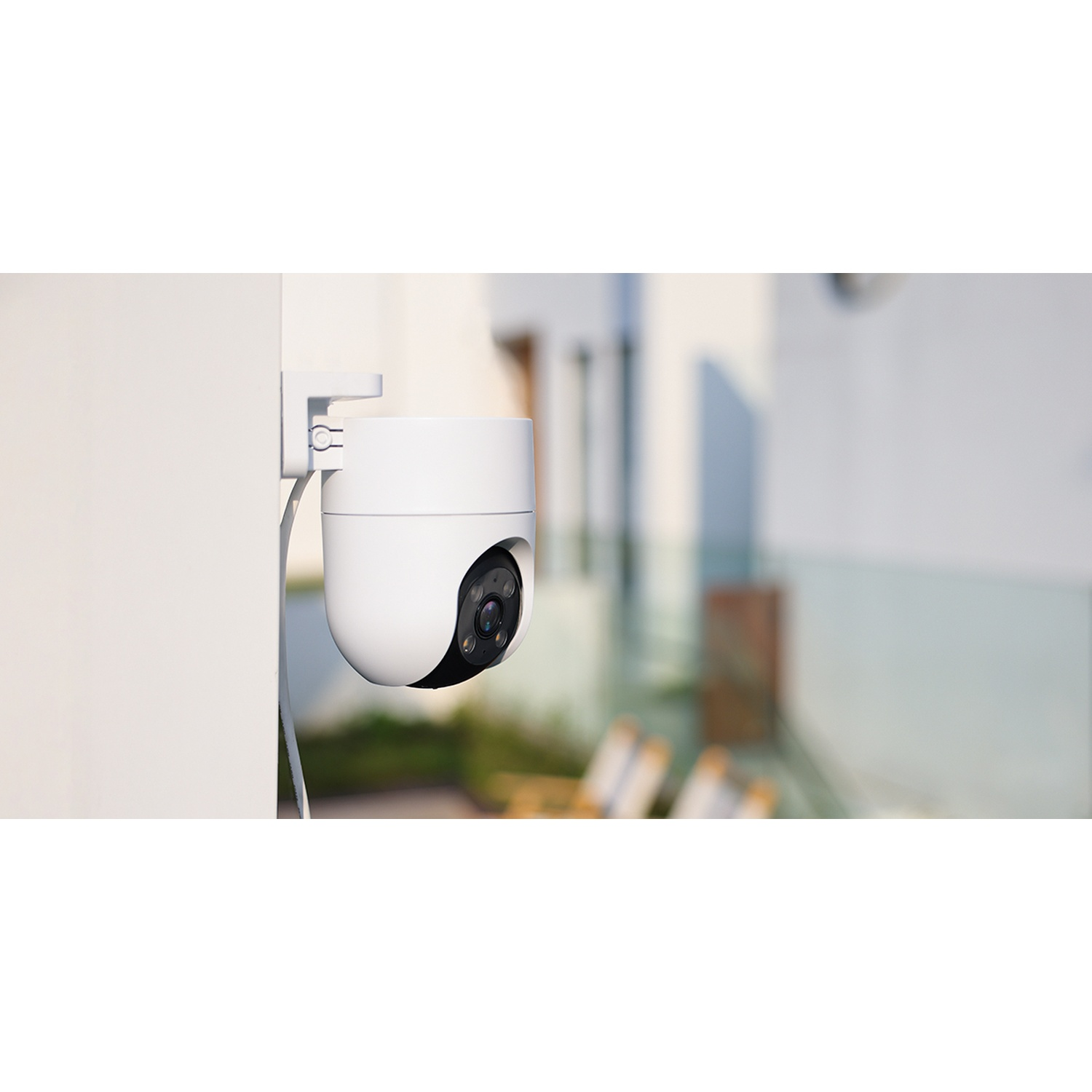 Una telecamera di sicurezza bianca montata su un muro bianco. La fotocamera è focalizzata sullo sfondo, che include una casa e alberi.