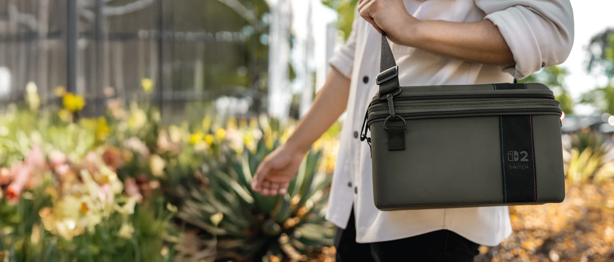 Personne tenant un sac vert avec une sangle noire. Le sac a un logo Nintendo Switch. Fleurs et plantes en arrière-plan.