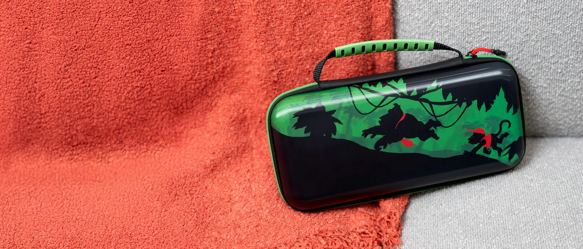 Una custodia nera per Nintendo Switch con un design a giungla verde e una maniglia verde è posizionata su una coperta rossa e una superficie grigia.