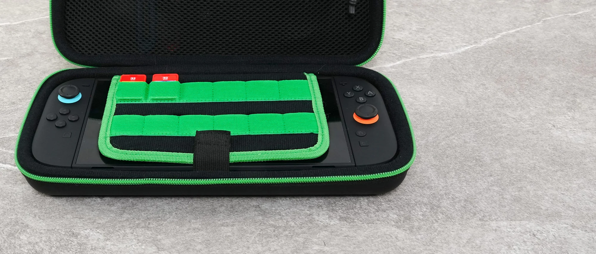 Custodia Switch nera e verde aperta con Switch all'interno. Area di stoccaggio verde per schede di gioco. Superficie grigia sullo sfondo.